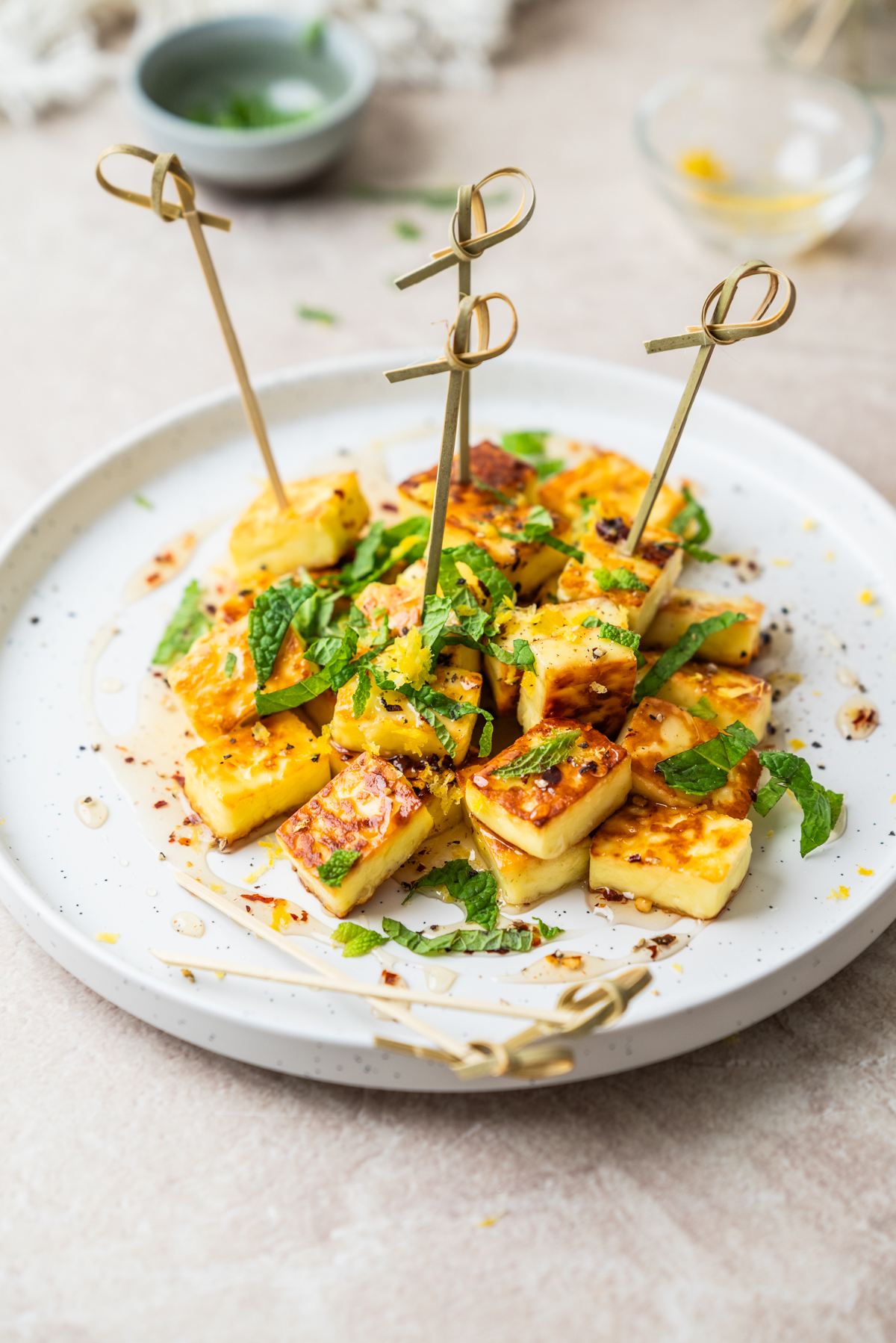 A plate of grilled halloumi cheese cubes on skewers, garnished with chopped mint, lemon zest, chili flakes, and a drizzle of honey.