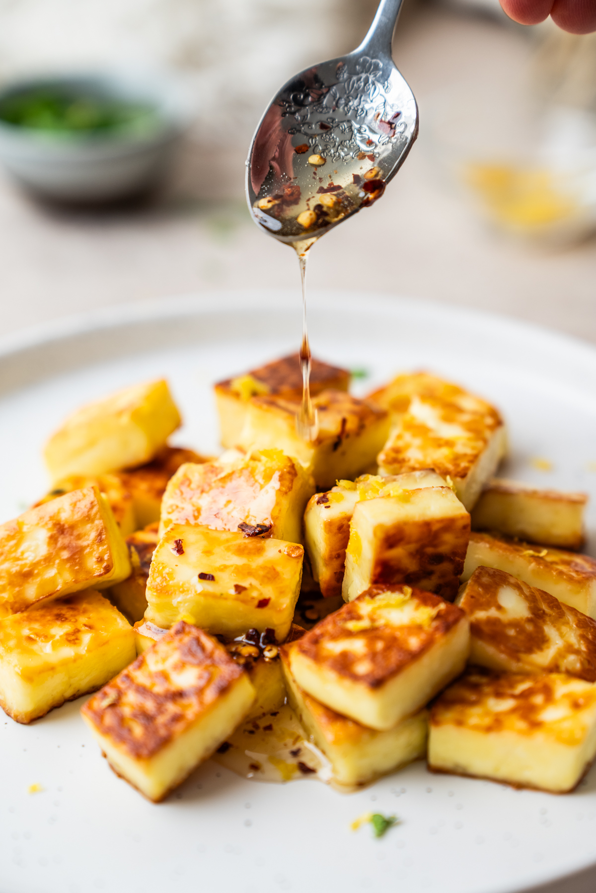 Golden-brown cubes of fried halloumi cheese on a white plate, with honey being drizzled from a spoon above.