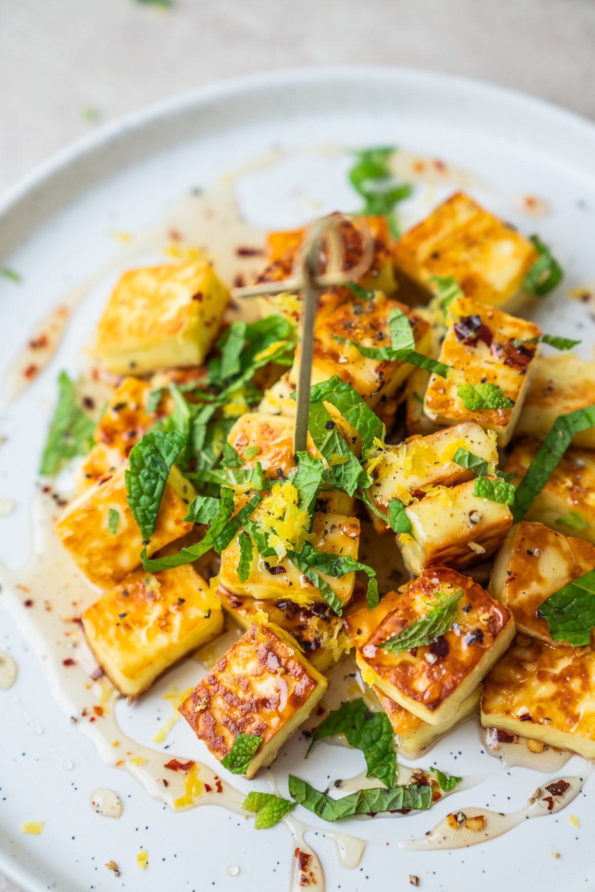A plate of golden-brown grilled halloumi cubes drizzled with honey, topped with chopped mint, lemon zest, and chili flakes, served on a white plate.