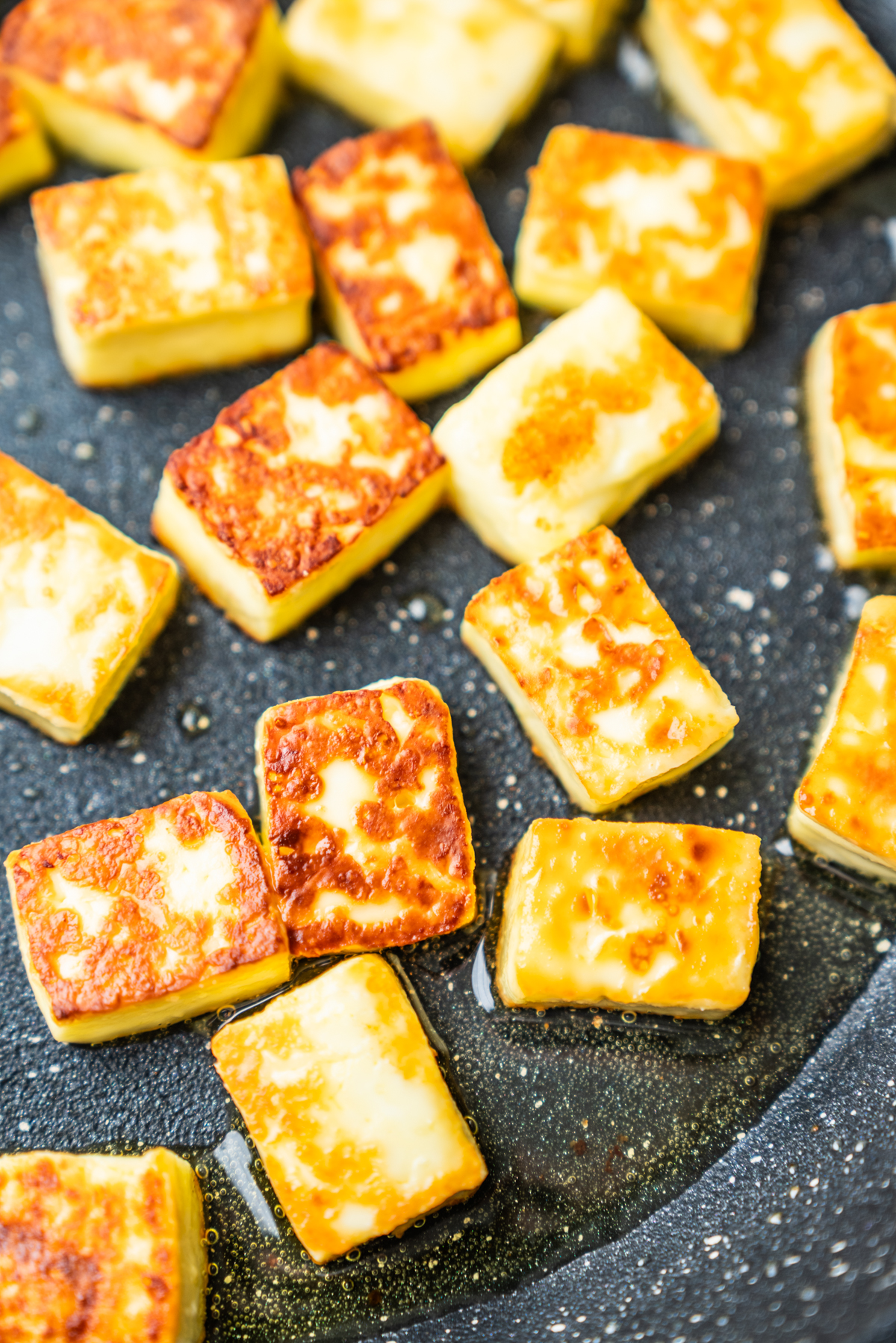 Golden-brown cubes of paneer cheese are sizzling in oil on a dark, speckled nonstick pan.