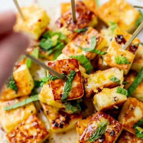 Cubes of grilled halloumi cheese garnished with chopped herbs, served on skewers on a white plate. A hand reaches in to pick up a skewer.