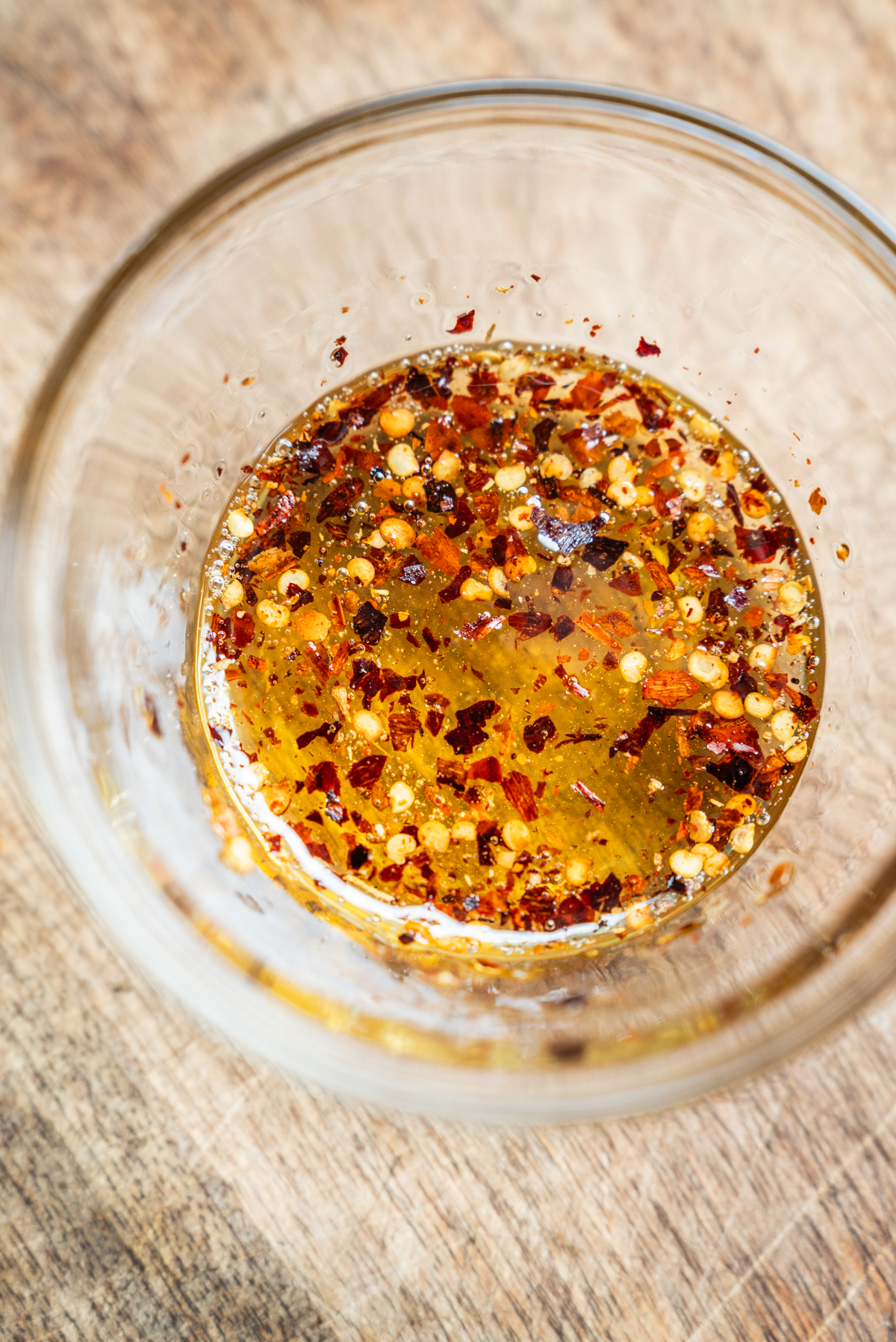 A close-up of a glass bowl filled with chili oil, showing red pepper flakes and seeds suspended in golden oil, placed on a wooden surface.