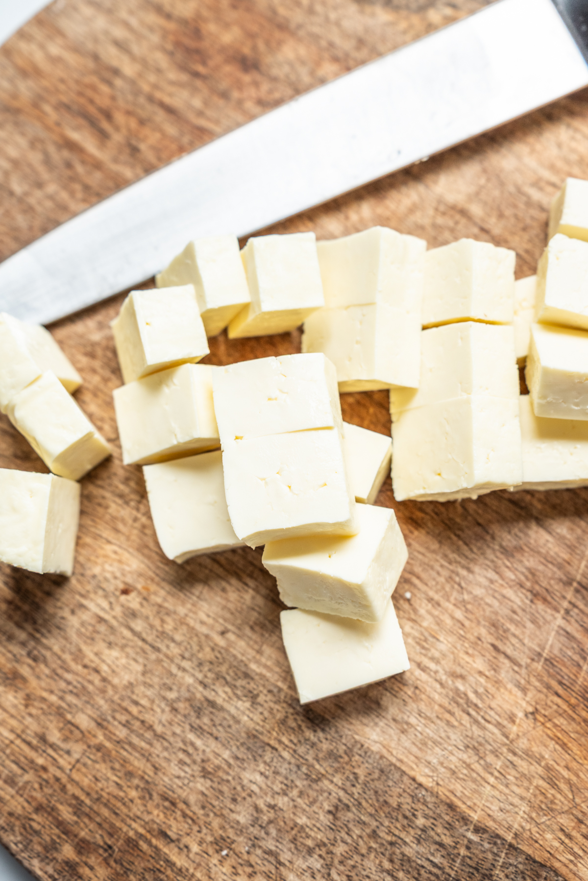 Cubes of white cheese are scattered on a wooden cutting board next to a large knife.