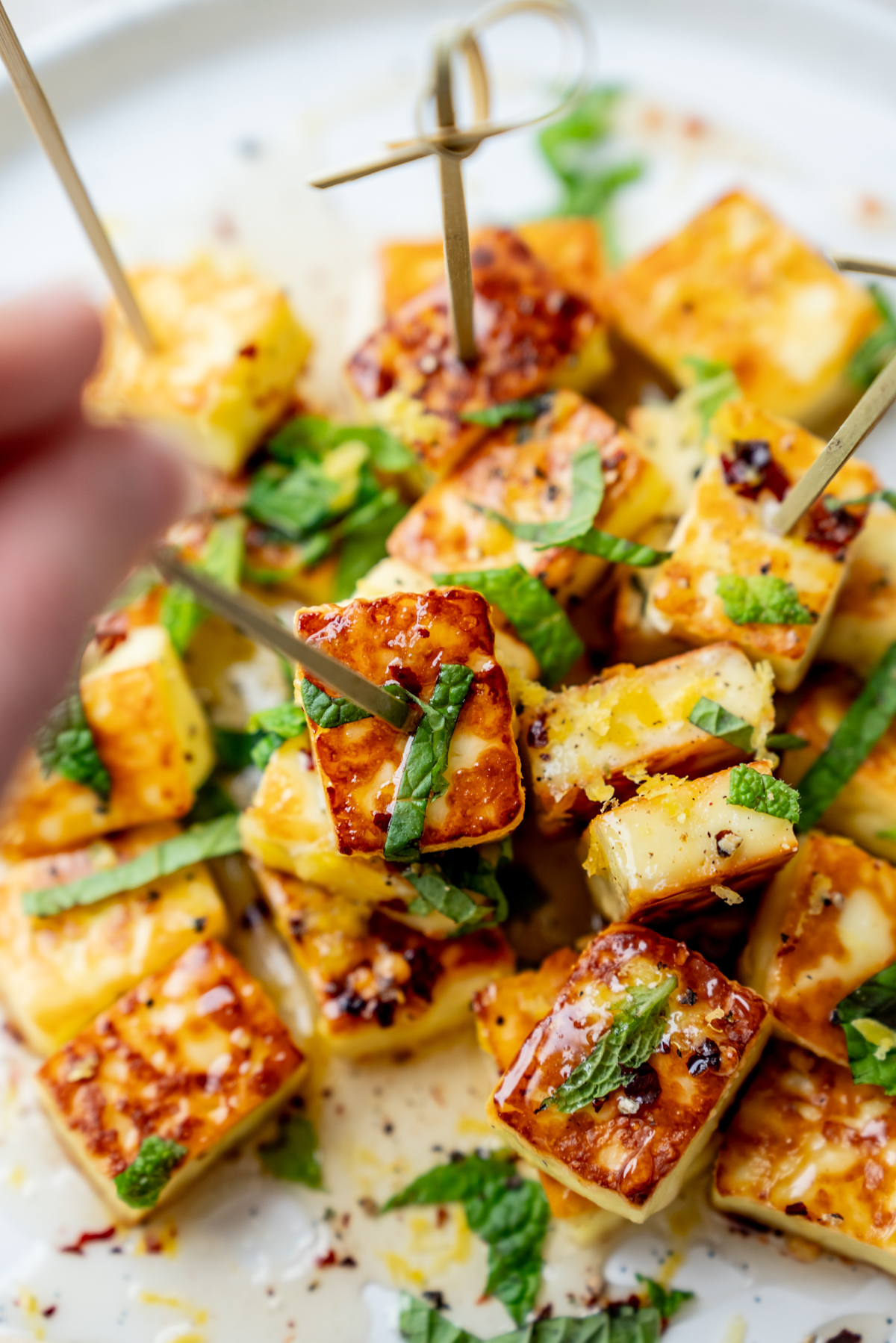 Cubes of grilled halloumi cheese garnished with chopped herbs, served on skewers on a white plate. A hand reaches in to pick up a skewer.