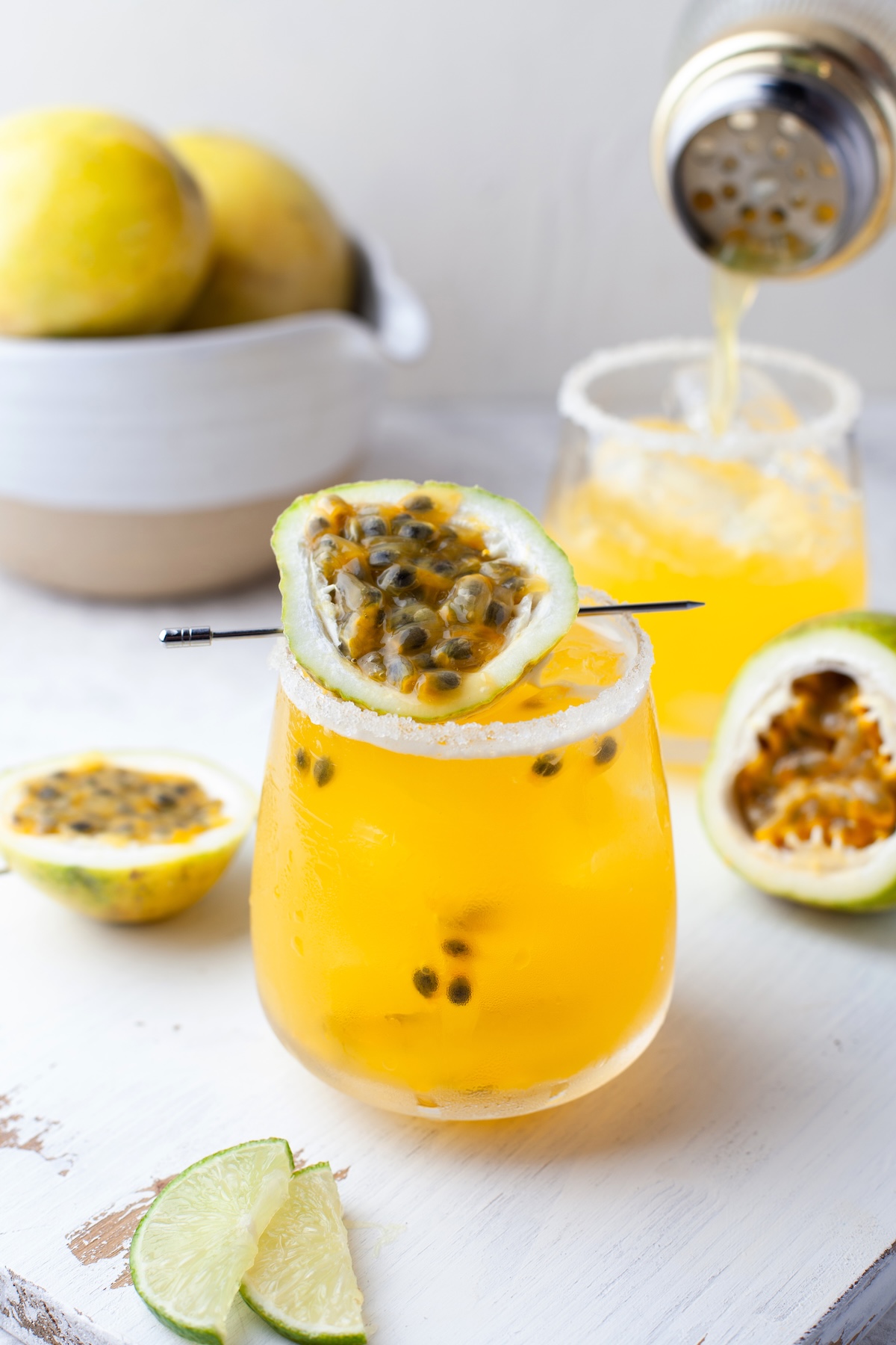 A glass of yellow passion fruit cocktail with passion fruit garnish, surrounded by lime slices and passion fruit halves, with a bowl of yellow fruits and a shaker pouring drink in the background.